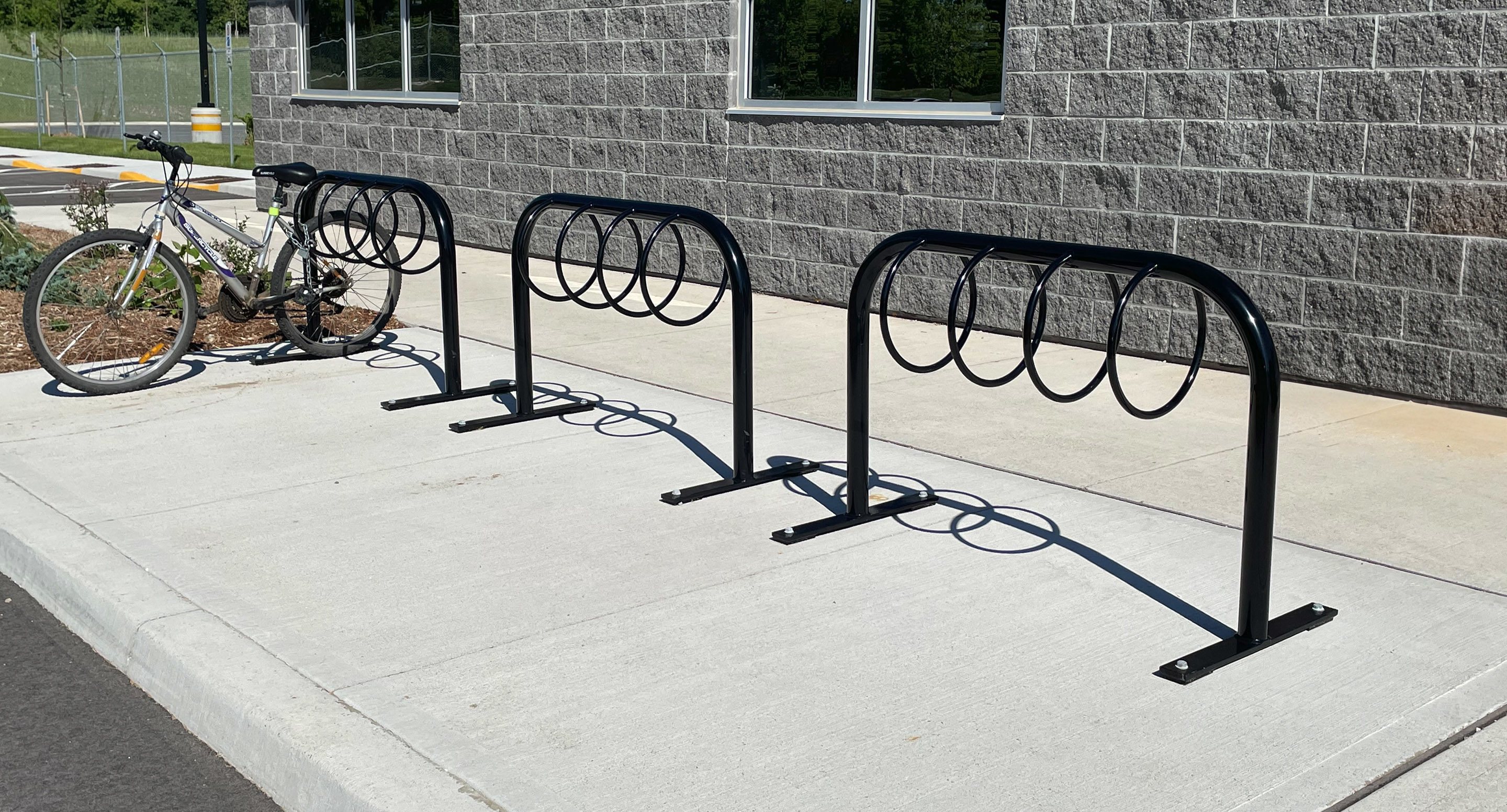 maglin_mbr-0300-bike-racks-3 A bike rack with circular slots is installed on a sidewalk near a gray brick building; one bicycle is parked and locked to the rack.