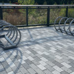 Two circular metal bike racks are positioned on a paved outdoor area with gray and white bricks, next to a black metal fence and plants in the background.