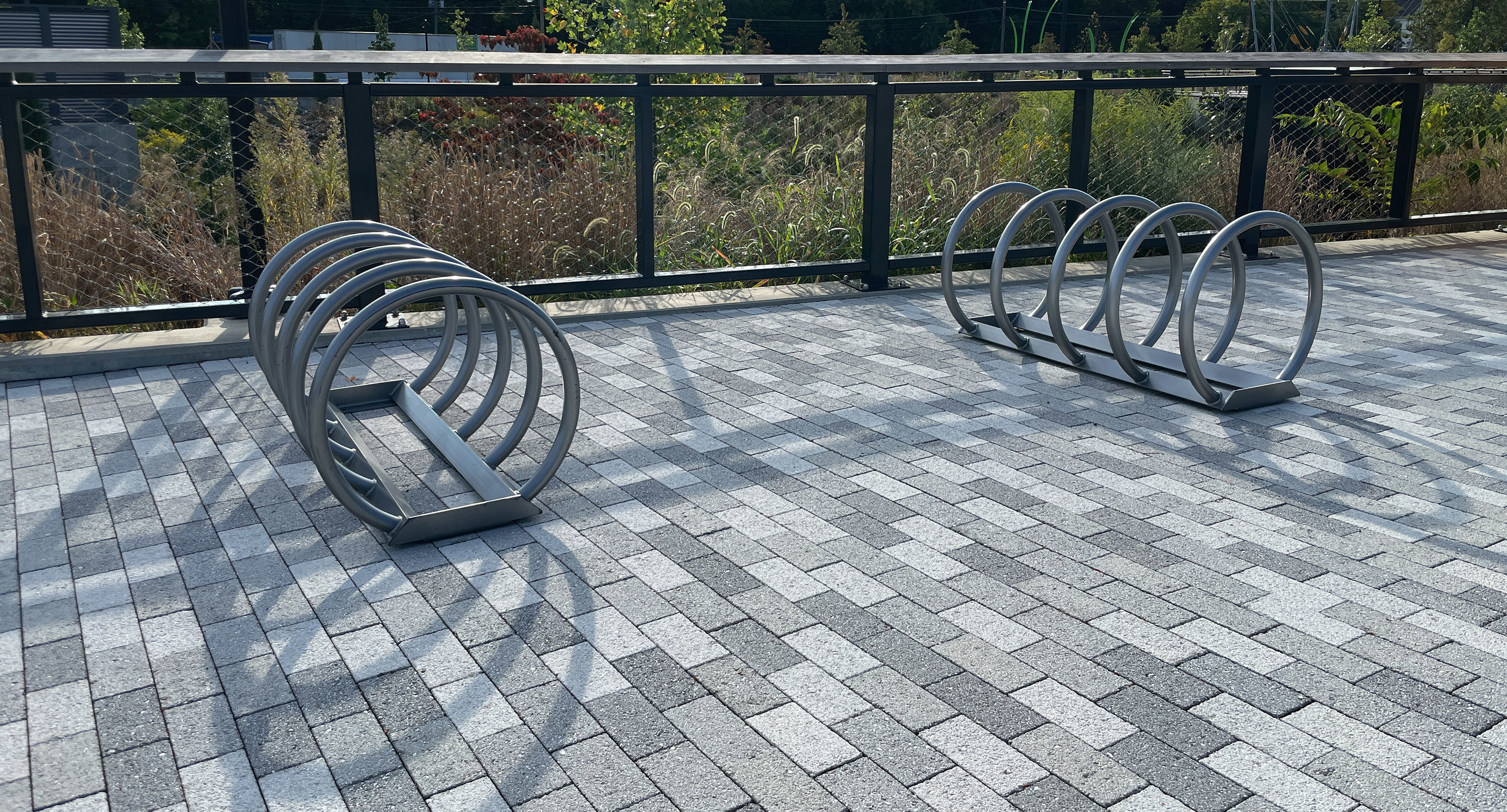 maglin_mbr-0350-bike-racks-1 Two circular metal bike racks are positioned on a paved outdoor area with gray and white bricks, next to a black metal fence and plants in the background.
