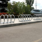 Two empty metal bike racks are installed on a sidewalk near a parking lot. A silver SUV is parked nearby, with trees and shrubs in the background.