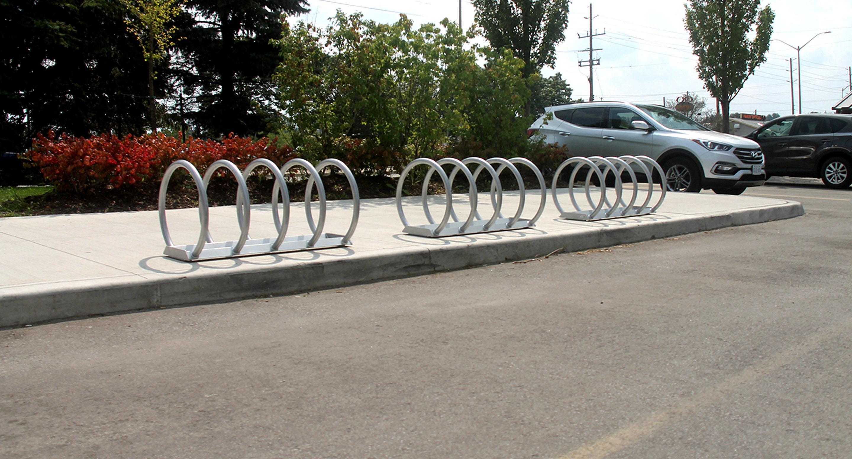 maglin_mbr-0350-bike-racks-2 Two empty metal bike racks are installed on a sidewalk near a parking lot. A silver SUV is parked nearby, with trees and shrubs in the background.