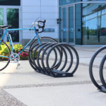 A single blue bicycle is parked in a circular metal bike rack outside a modern glass building, with flowers and greenery nearby.