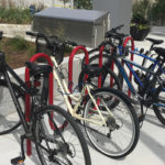 Four bicycles of different colors are parked and locked to red bike racks on a concrete sidewalk near landscaping and a building.