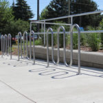 A bike rack with empty metal loops is installed on a concrete sidewalk near a landscaped area with greenery and trees.