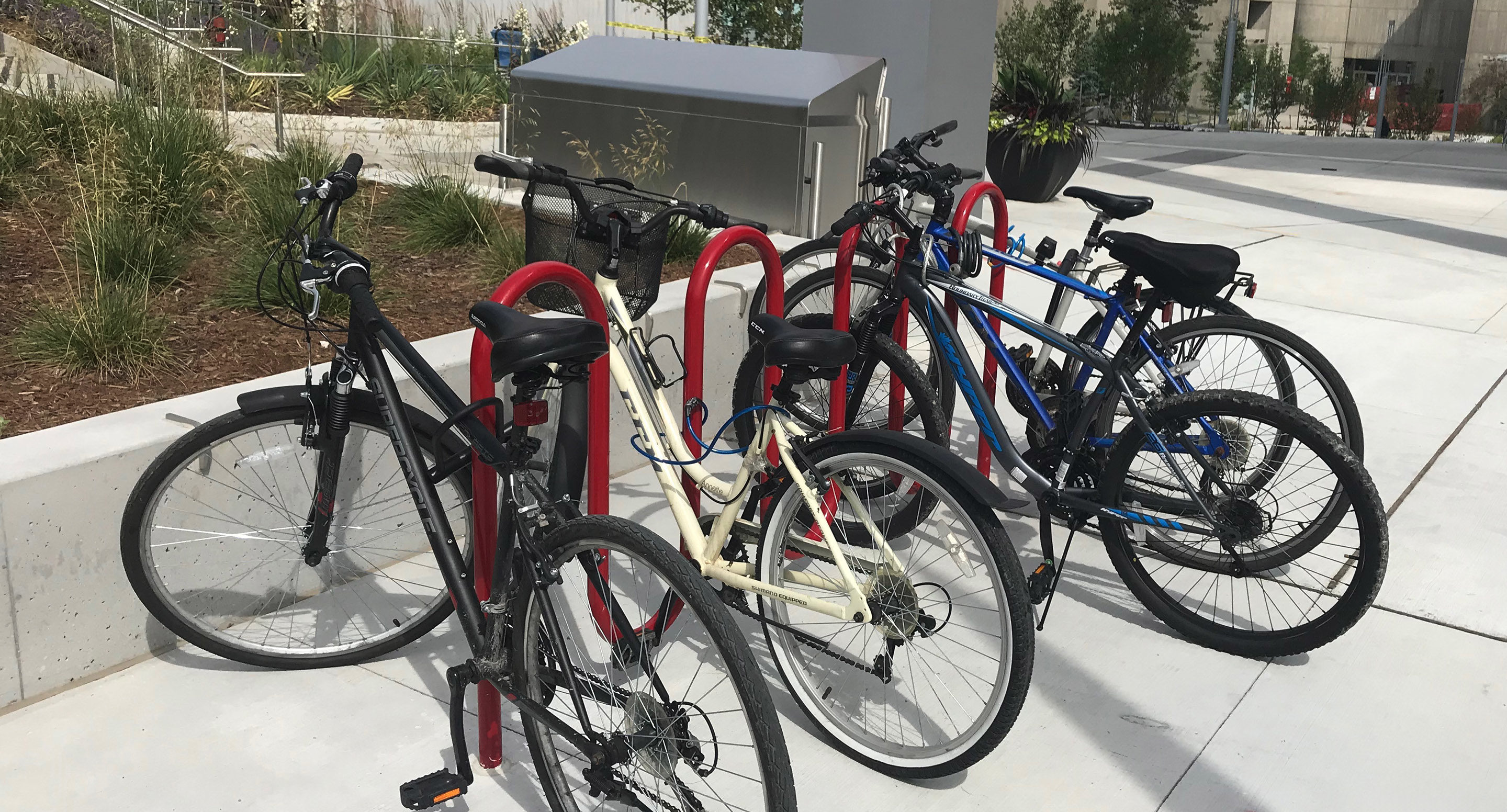 maglin_mbr-0400-bike-racks-1 Four bicycles of different colors are parked and locked to red bike racks on a concrete sidewalk near landscaping and a building.