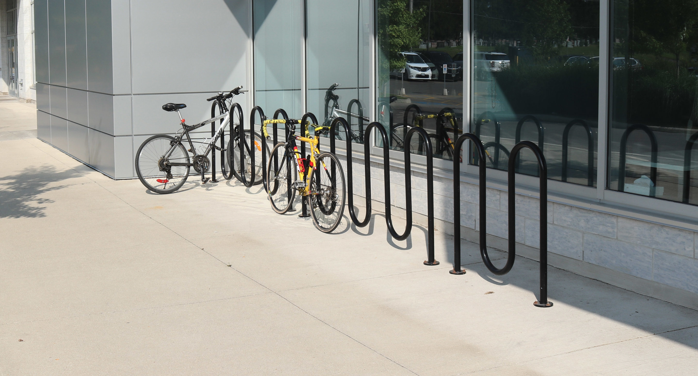 maglin_mbr-0400-bike-racks-2 Several bicycles are parked and locked to black metal bike racks outside a modern building with large windows.