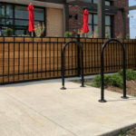 Outdoor restaurant patio with wooden planters, black metal fencing, two red umbrellas, and a bike rack on a concrete sidewalk.