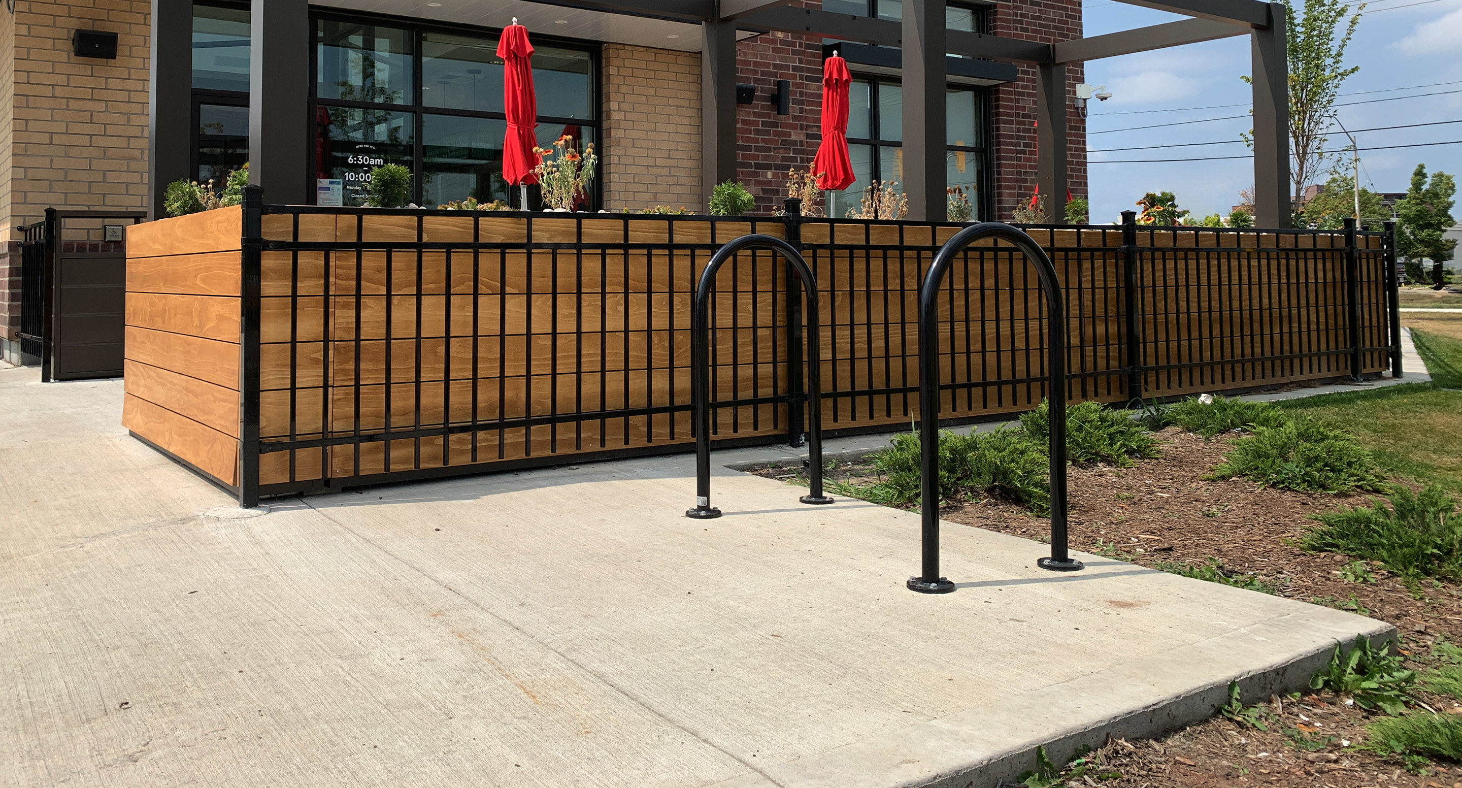 maglin_mbr-0500-bike-racks-1 Outdoor restaurant patio with wooden planters, black metal fencing, two red umbrellas, and a bike rack on a concrete sidewalk.