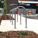 A row of metal bike racks is installed on a sidewalk next to a landscaped area outside a commercial building.