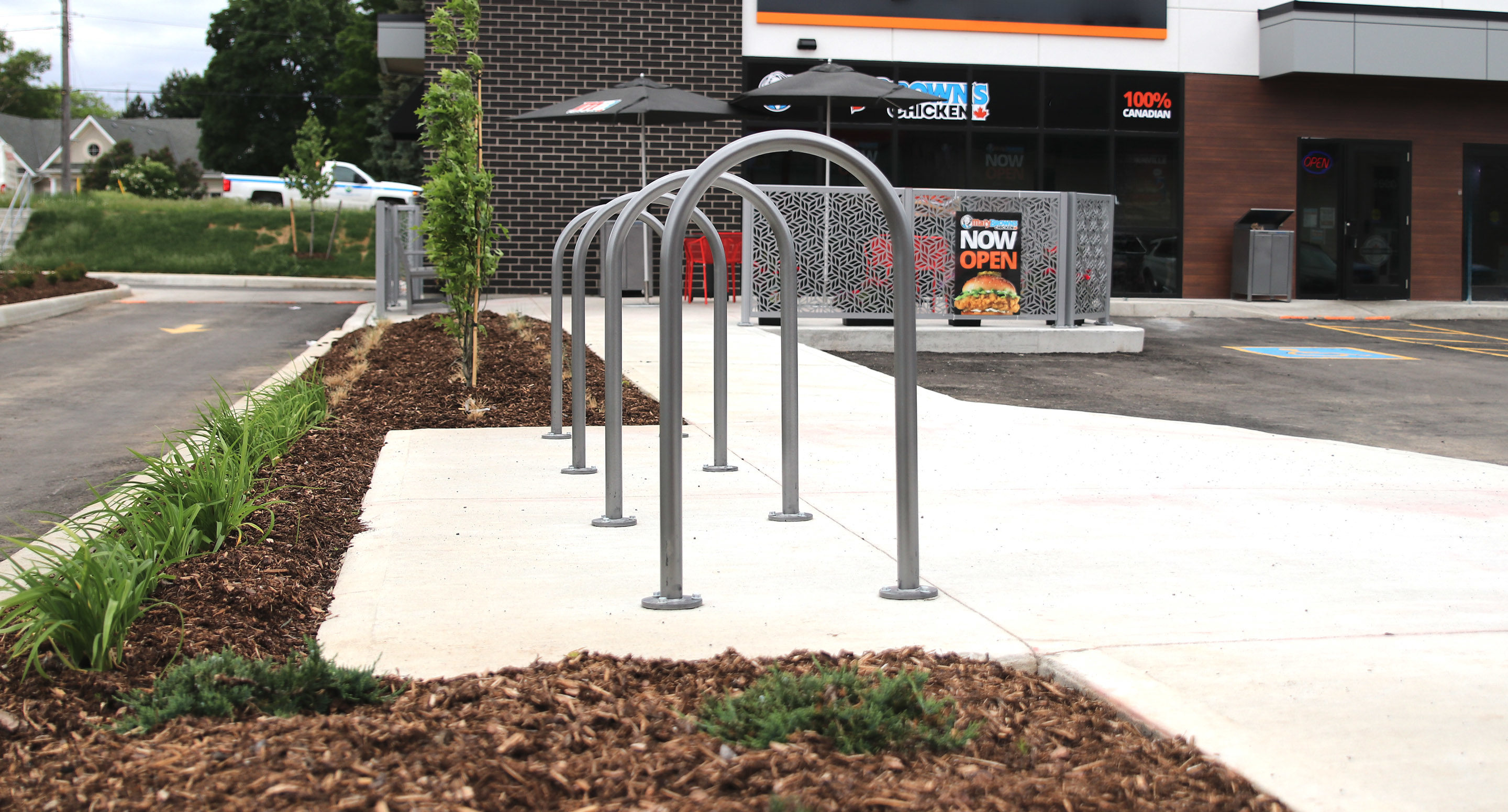 maglin_mbr-0500-bike-racks-2 A row of metal bike racks is installed on a sidewalk next to a landscaped area outside a commercial building.