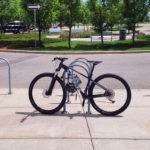 A black bicycle is parked and locked to a bike rack on a sunny sidewalk near a street with trees and parked cars in the background.