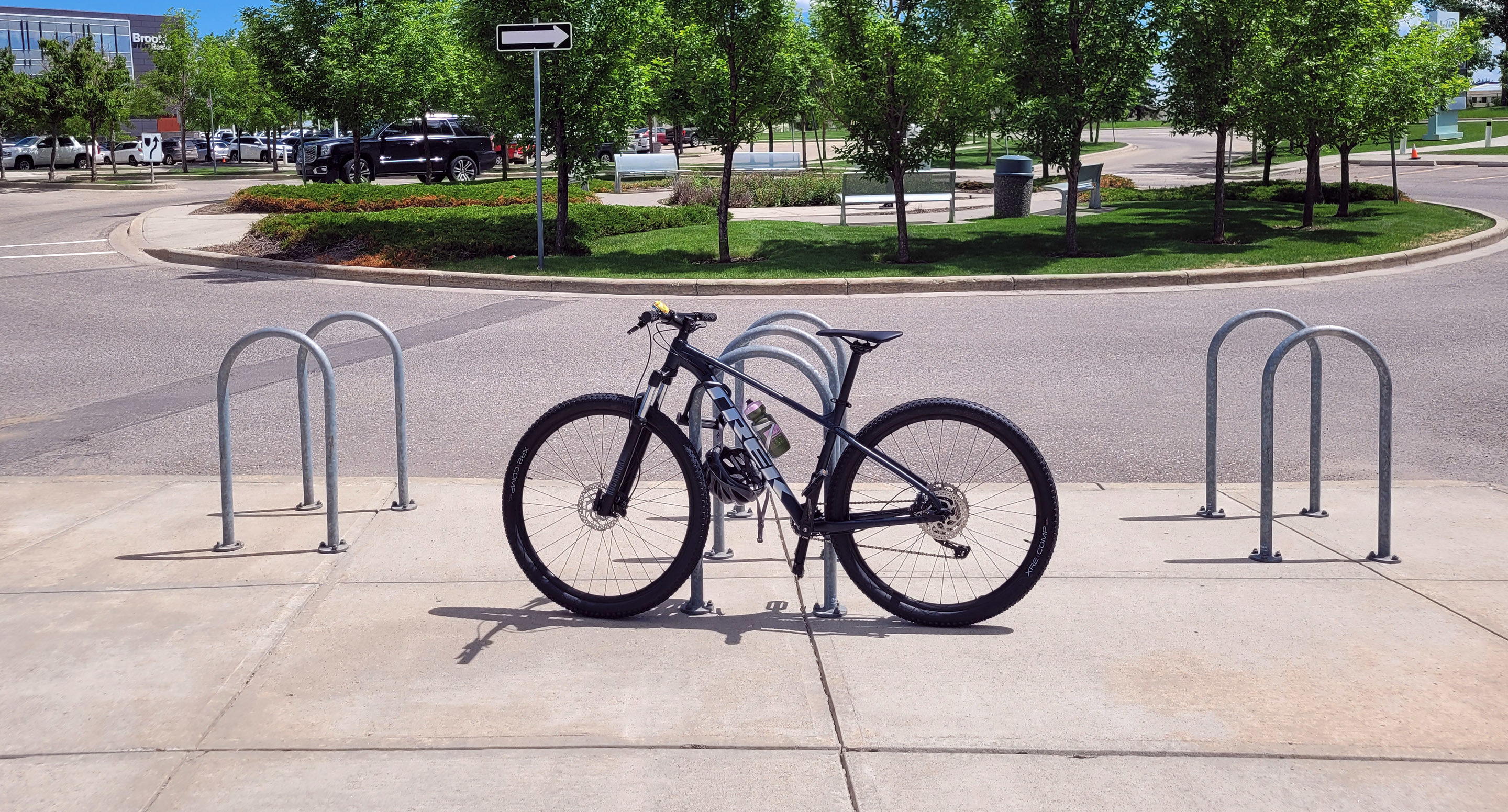 maglin_mbr-0500-bike-racks-3 A black bicycle is parked and locked to a bike rack on a sunny sidewalk near a street with trees and parked cars in the background.