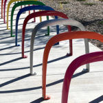 Des supports à vélos métalliques colorés en forme d'arcs sont installés en rangée le long d'un trottoir, projetant des ombres courbes sur le béton en plein soleil.