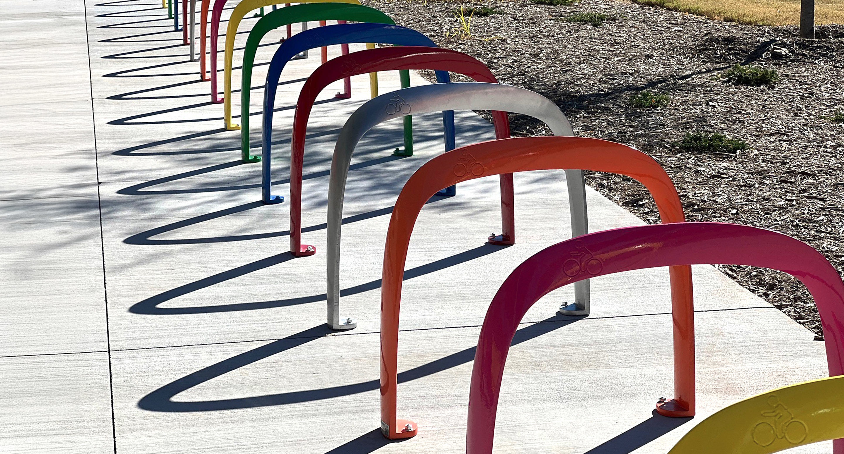 Des supports à vélos métalliques colorés en forme d'arcs sont installés en rangée le long d'un trottoir, projetant des ombres courbes sur le béton en plein soleil.