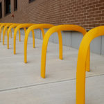 Une rangée de supports à vélos en métal jaune est alignée sur un trottoir en béton à côté d'un bâtiment en briques.