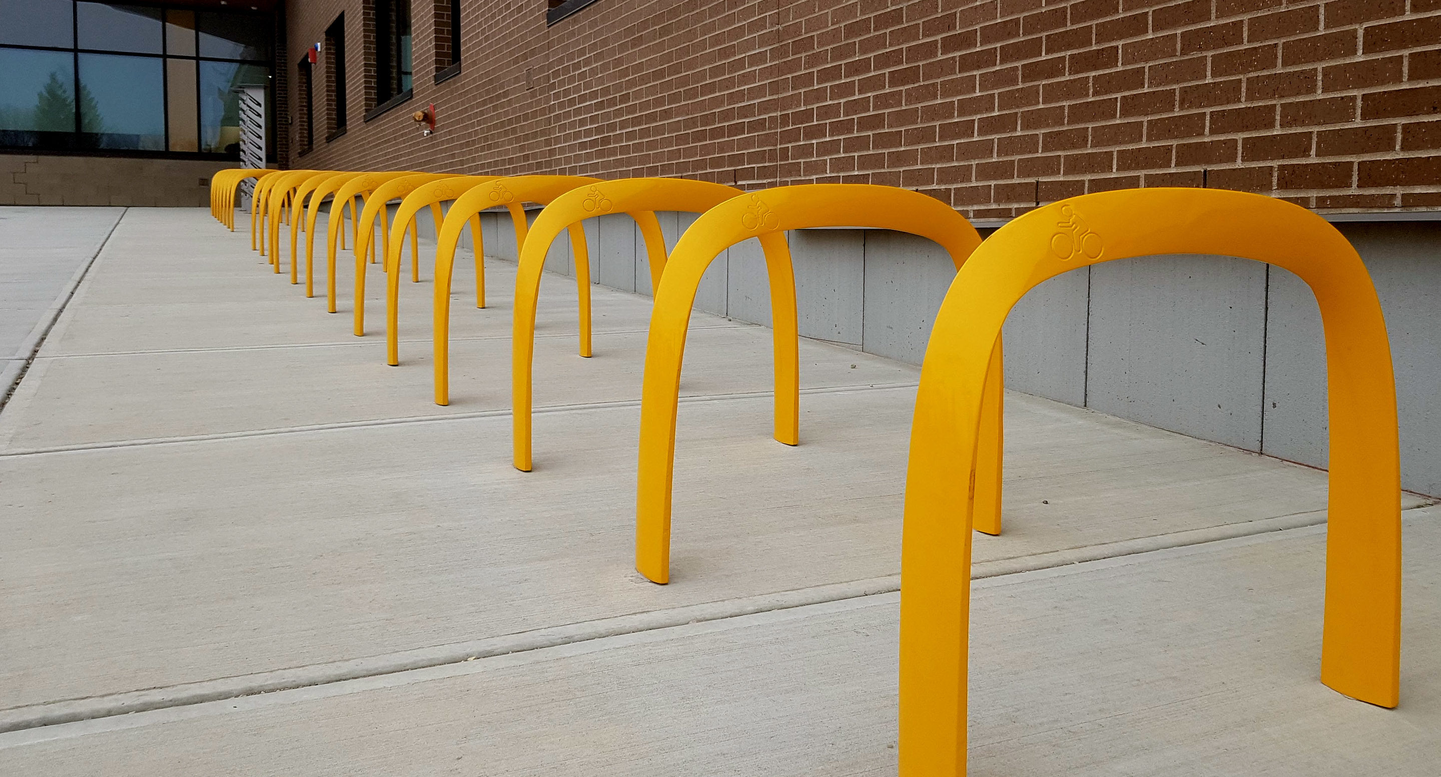 maglin_mbr-1600-bike-racks-2 Une rangée de supports à vélos en métal jaune est alignée sur un trottoir en béton à côté d'un bâtiment en briques.