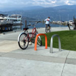 Un vélo rouge est garé sur un support à vélos près d'un port de plaisance, avec des bateaux, une sculpture et un cycliste en arrière-plan.