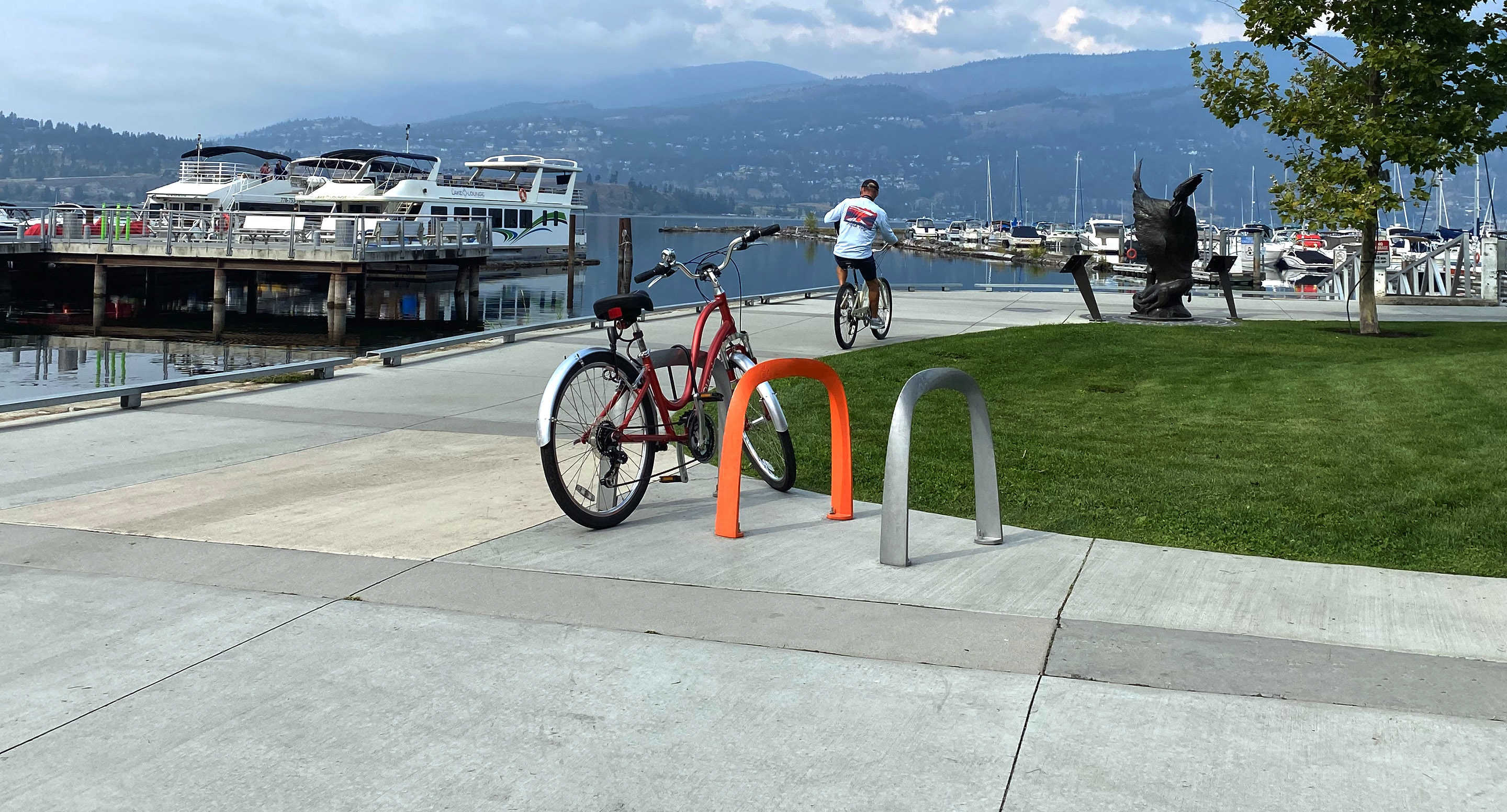 Un vélo rouge est garé sur un support à vélos près d'un port de plaisance, avec des bateaux, une sculpture et un cycliste en arrière-plan.