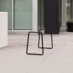 Two black metal bike racks are installed on a wide concrete sidewalk beside modern buildings with large windows and rectangular planters.