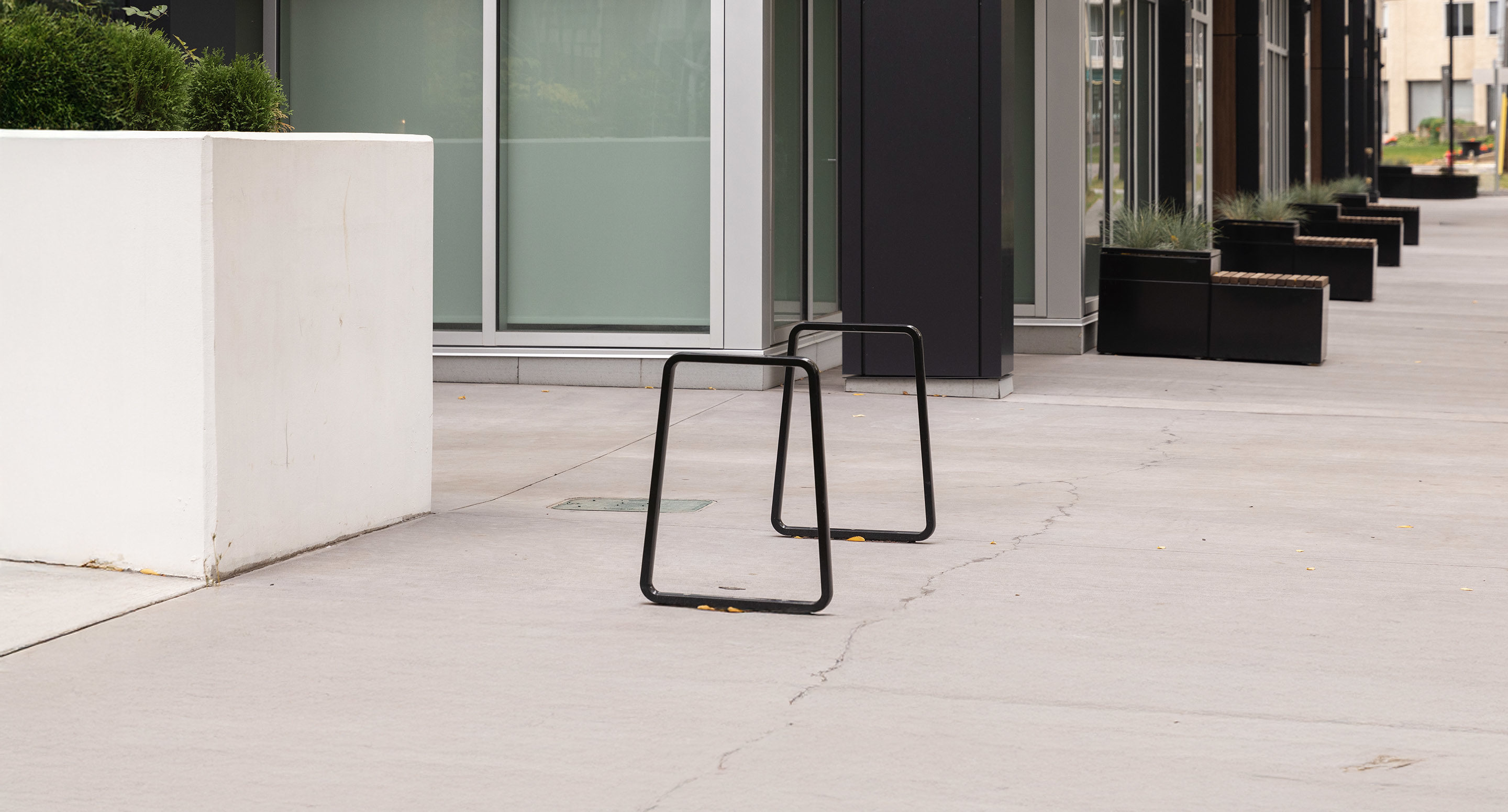 maglin_mbr-2300-bike-racks-1 Two black metal bike racks are installed on a wide concrete sidewalk beside modern buildings with large windows and rectangular planters.