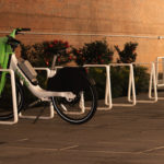 A single green and white rental bike is parked at an otherwise empty bike rack along a paved sidewalk next to a brick wall and some shrubs.