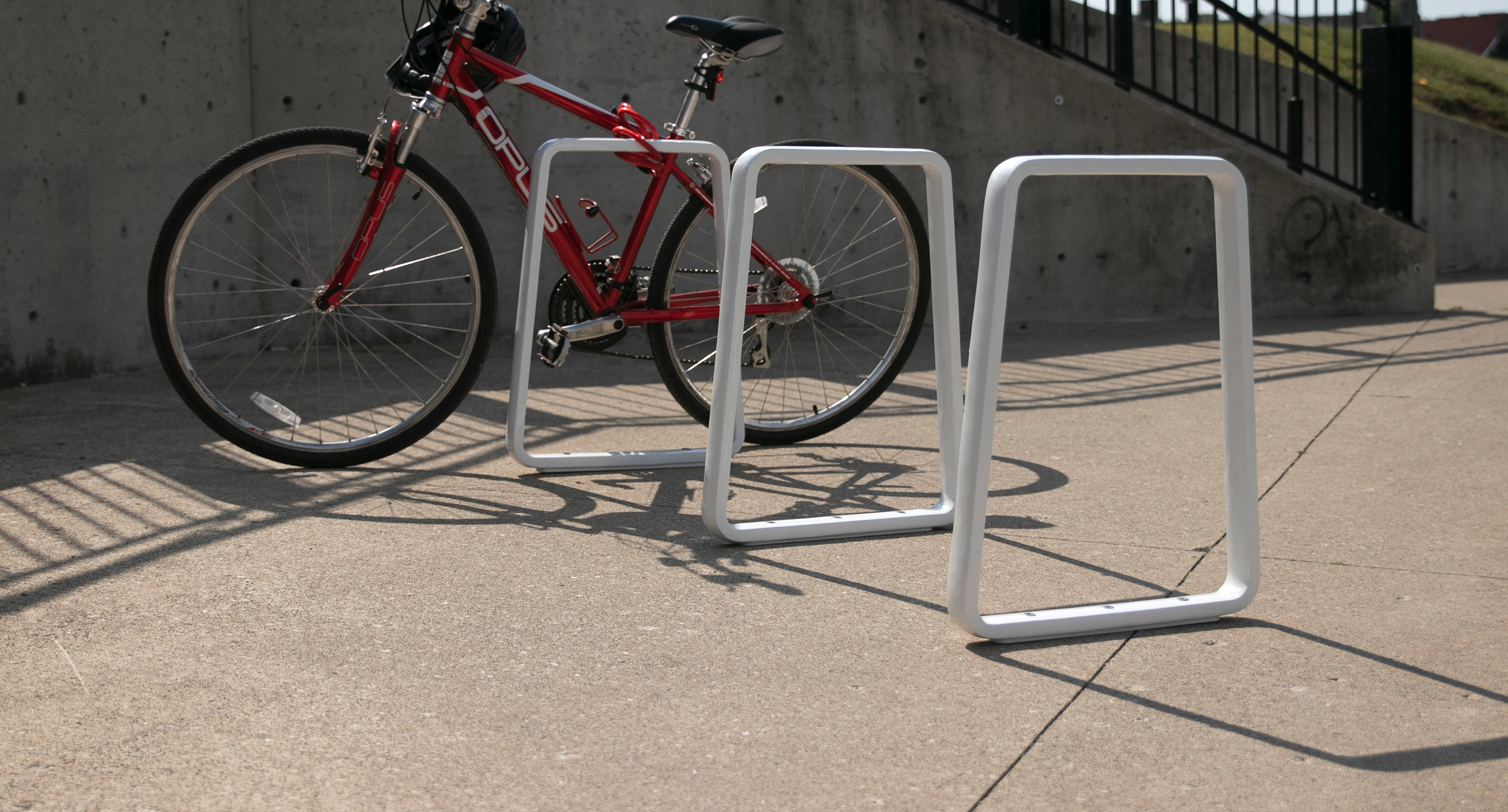 maglin_mbr-2300-bike-racks-3 A red bicycle is locked to a white metal bike rack on a concrete sidewalk next to a stairway with a black handrail.