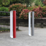 Two modern, upright bike racks—one red, one white—installed on a paved area next to black railings and landscaped bushes in a park setting.
