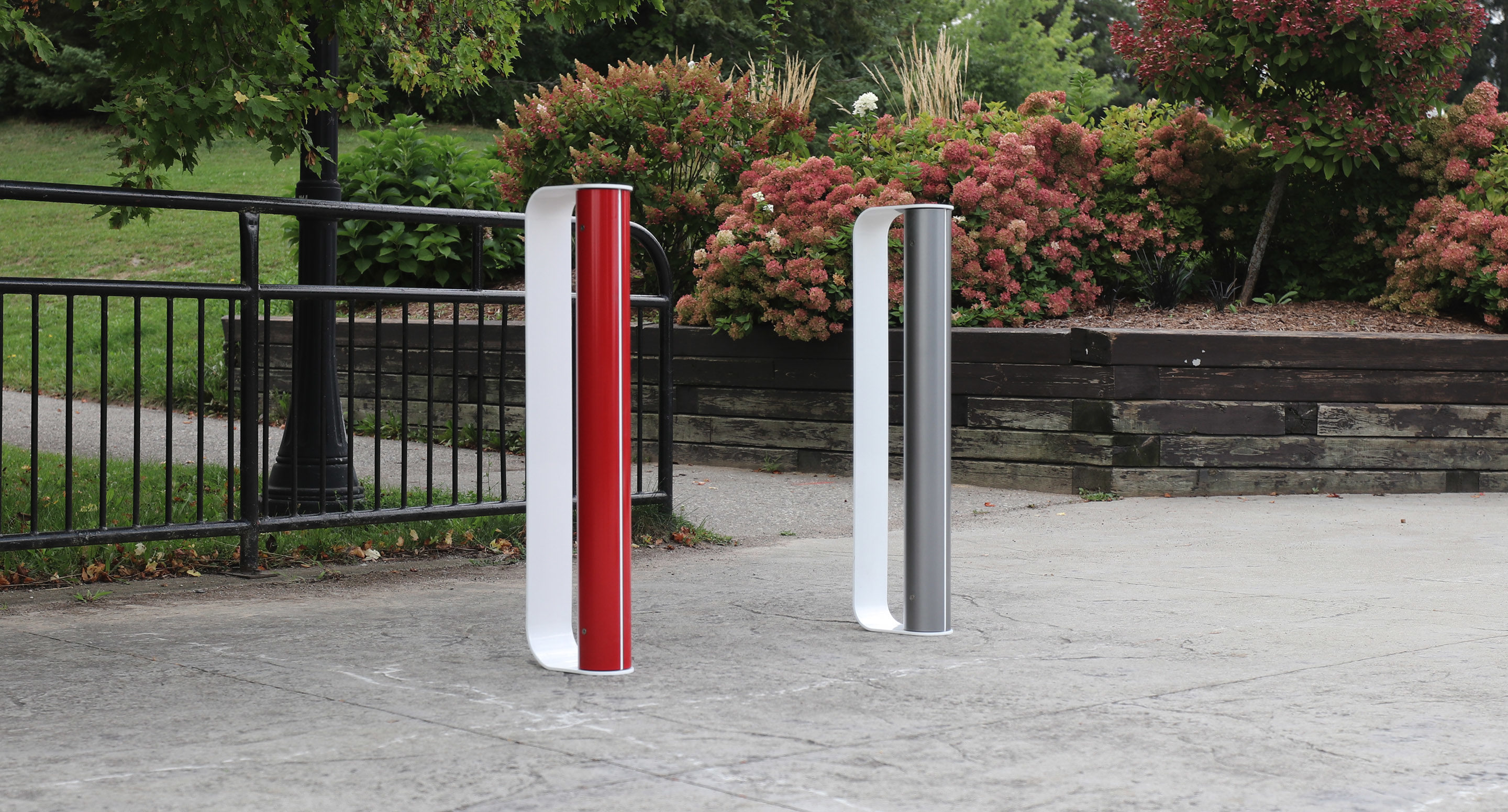 maglin_mbr-2900-bike-racks-1 Two modern, upright bike racks—one red, one white—installed on a paved area next to black railings and landscaped bushes in a park setting.