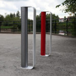 Two modern, vertical metal bike racks—one gray, one red—stand on a concrete surface outdoors near a black metal fence and greenery.