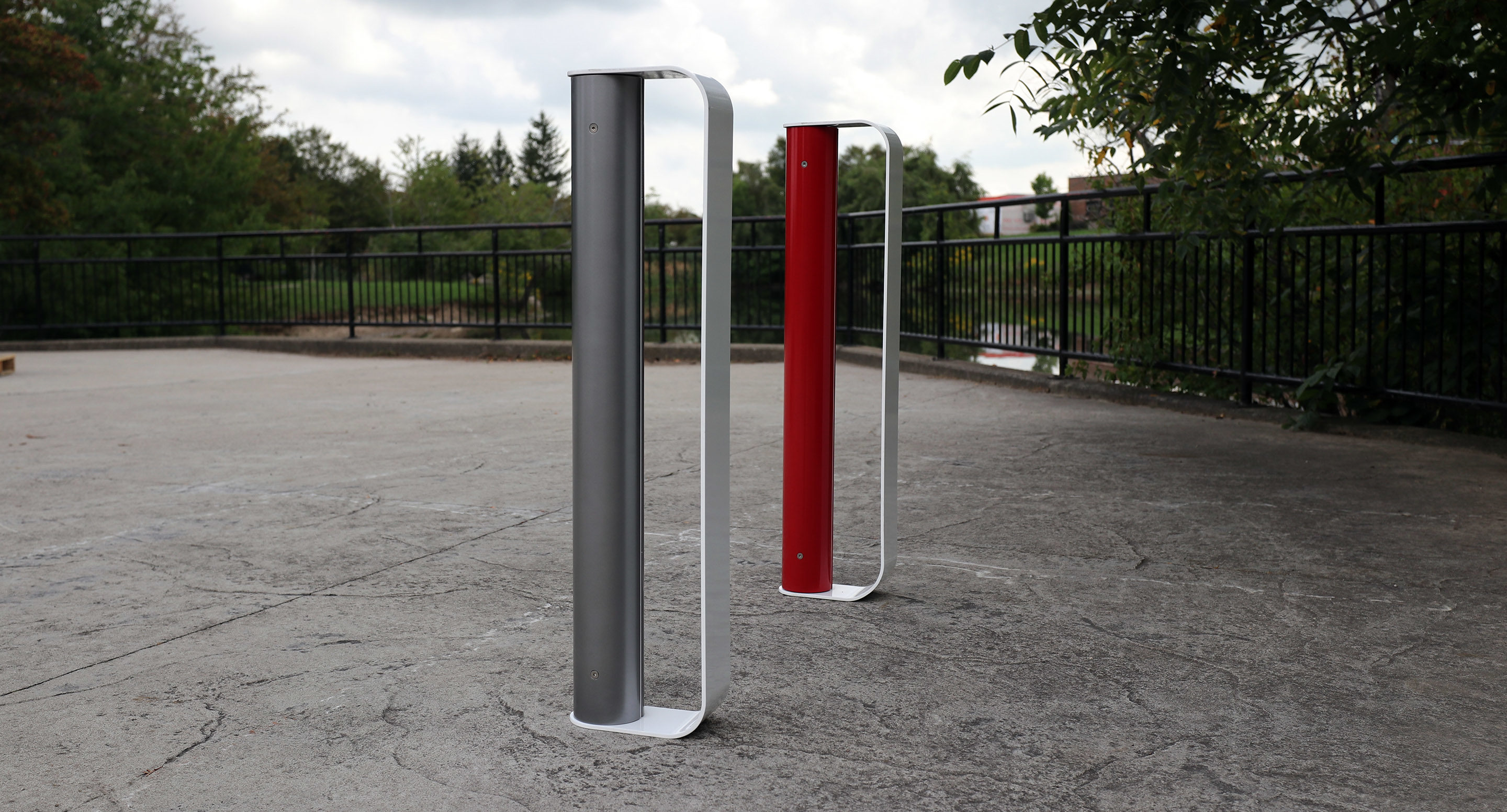 maglin_mbr-2900-bike-racks-2 Two modern, vertical metal bike racks—one gray, one red—stand on a concrete surface outdoors near a black metal fence and greenery.