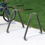 A blue bicycle is parked in a metal bike rack on a concrete walkway next to a grassy lawn with small plants and mulch.