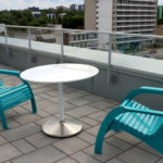 Two turquoise patio chairs and a round white table on a rooftop terrace with a cityscape in the background.