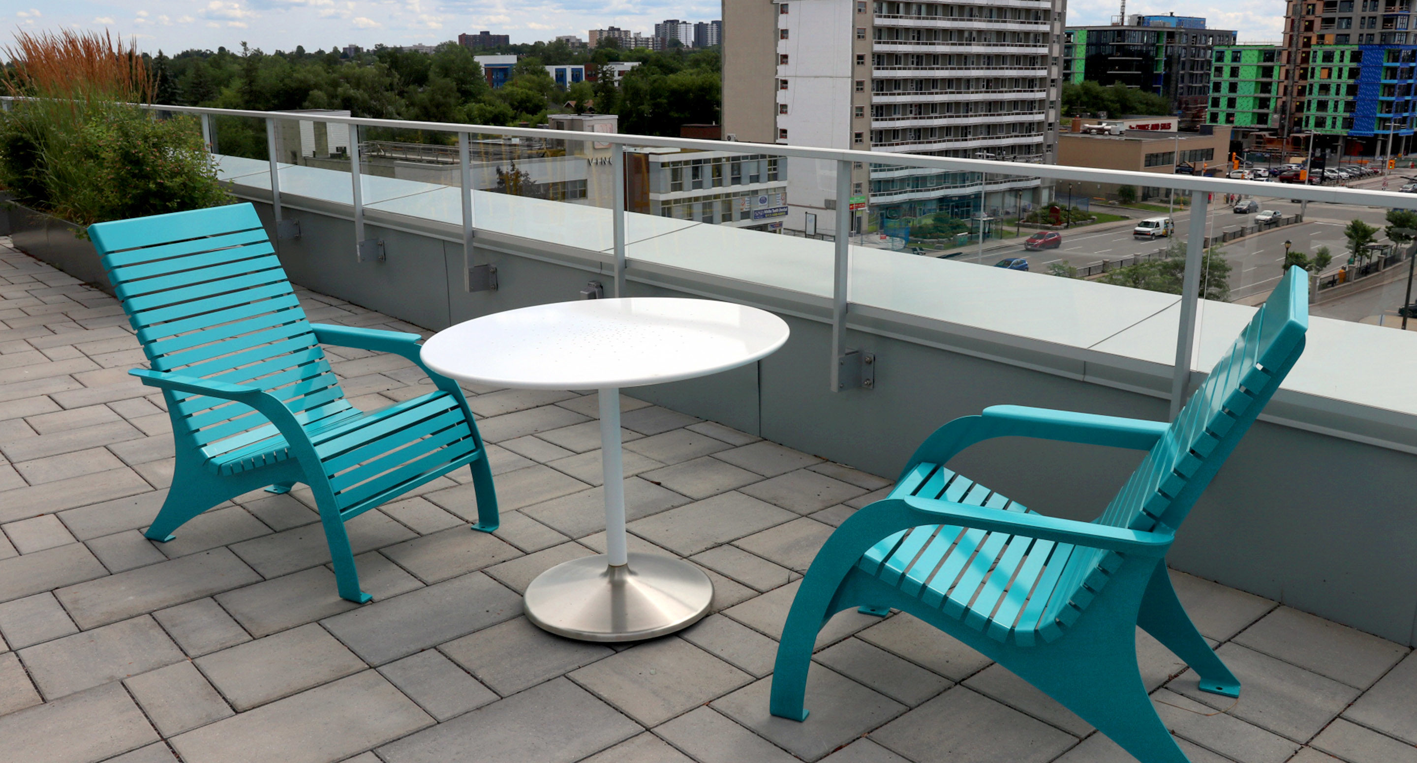 maglin_mch-0720-chairs-1 Two turquoise patio chairs and a round white table on a rooftop terrace with a cityscape in the background.