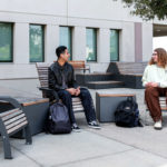 Two students sit on outdoor benches with backpacks at their feet, conversing outside a building with large windows and trees nearby.