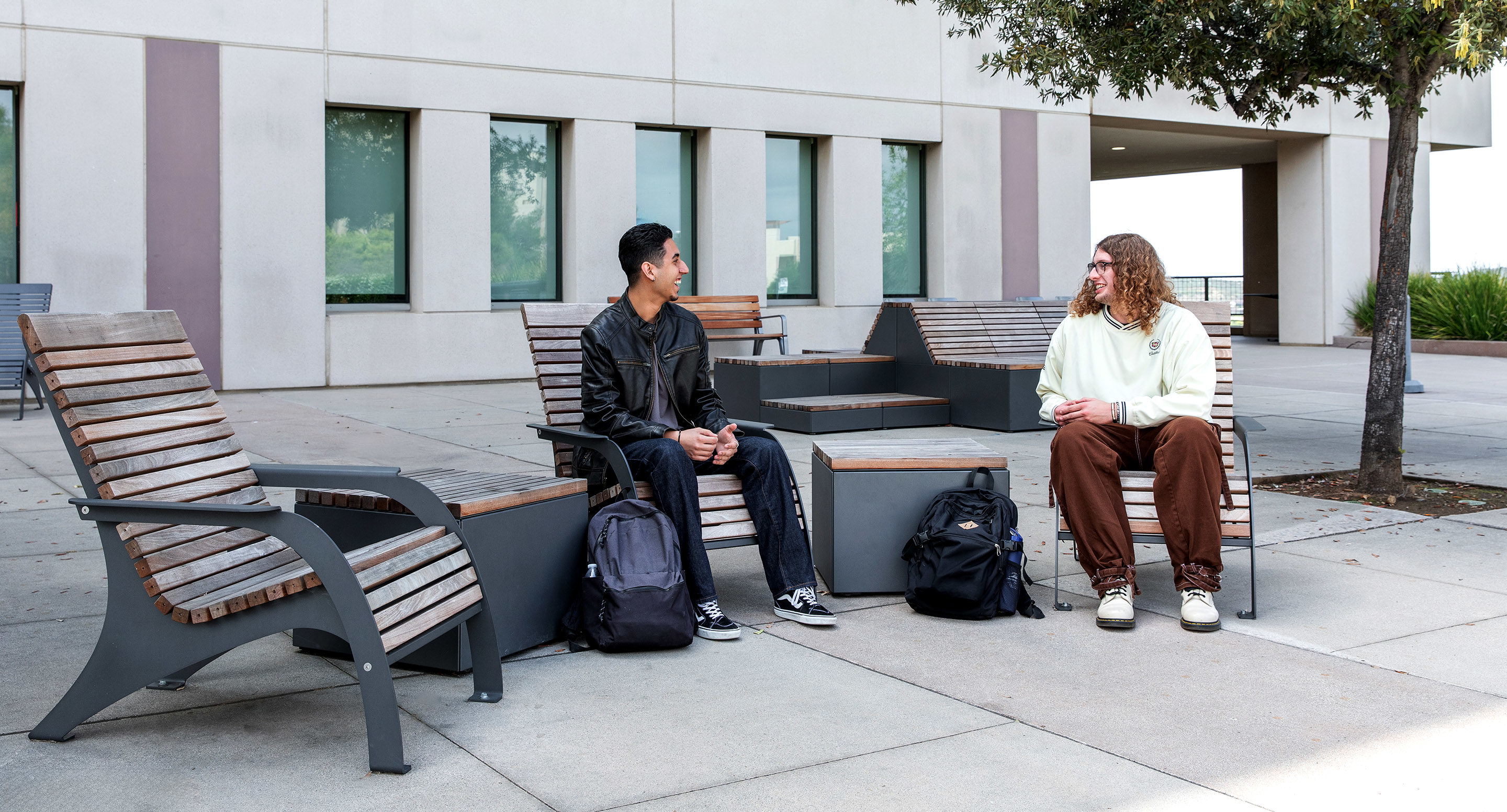 Two students sit on outdoor benches with backpacks at their feet, conversing outside a building with large windows and trees nearby.