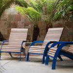 Three wooden lounge chairs with blue metal frames are arranged in a row on a patio, surrounded by lush ferns and greenery against a stone tile wall.