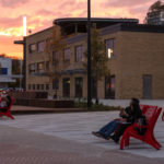 People sit on red chairs in an open urban plaza at sunset, with a modern brick building and trees in the background.