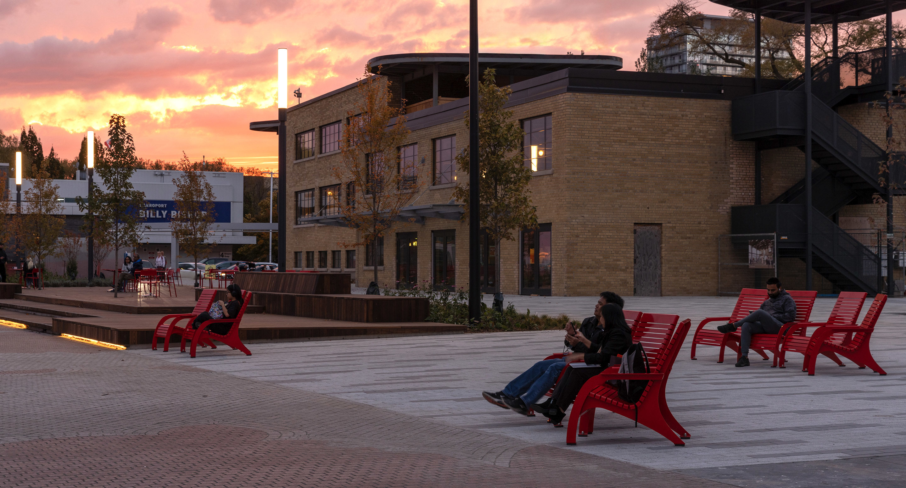 People sit on red chairs in an open urban plaza at sunset, with a modern brick building and trees in the background.