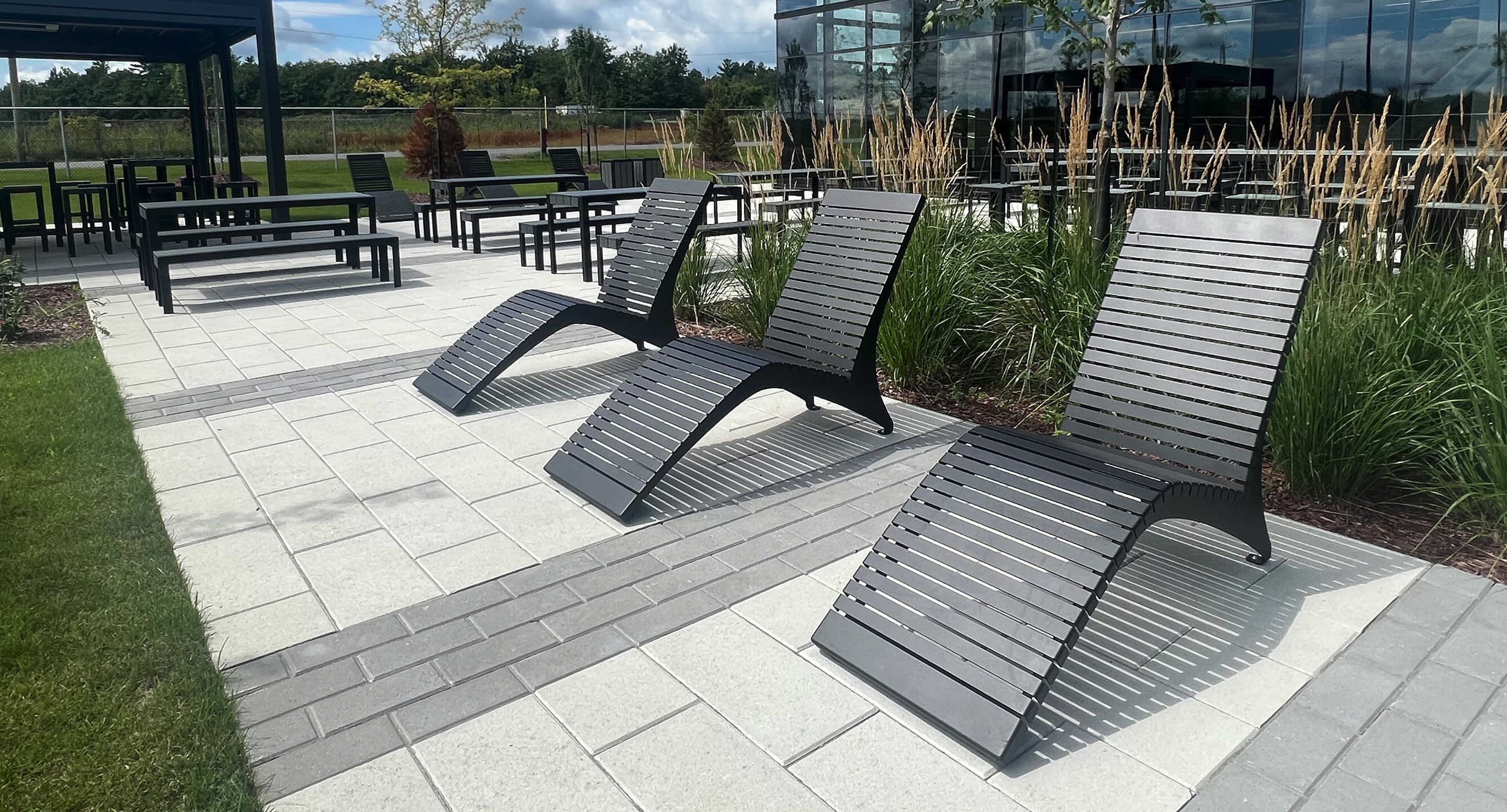 Three black lounge chairs and several tables and chairs are arranged on a paved outdoor patio next to tall grasses and a modern glass building.