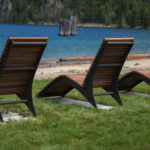Three wooden lounge chairs are positioned on grass facing a lake, with a sandy shore and forested hills in the background.