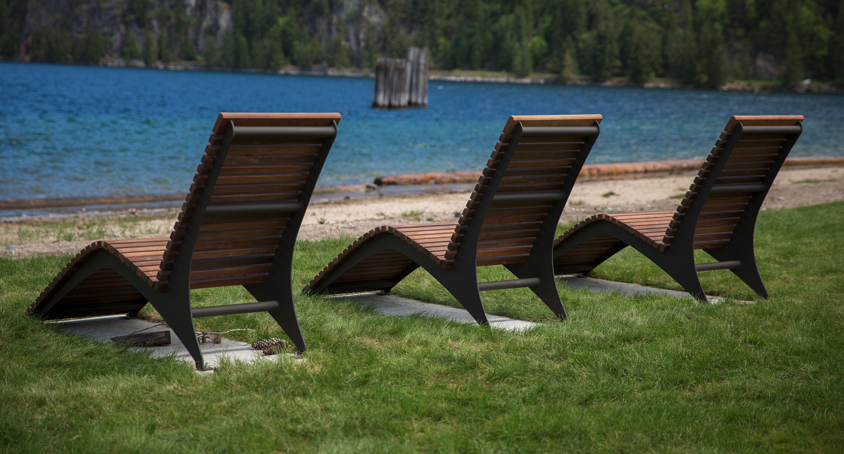 maglin_mch-0720-chaise-lounge-2 Three wooden lounge chairs are positioned on grass facing a lake, with a sandy shore and forested hills in the background.