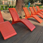 Six red slatted lounge chairs are arranged in a row on a wooden deck, with trees and green chairs visible in the background.