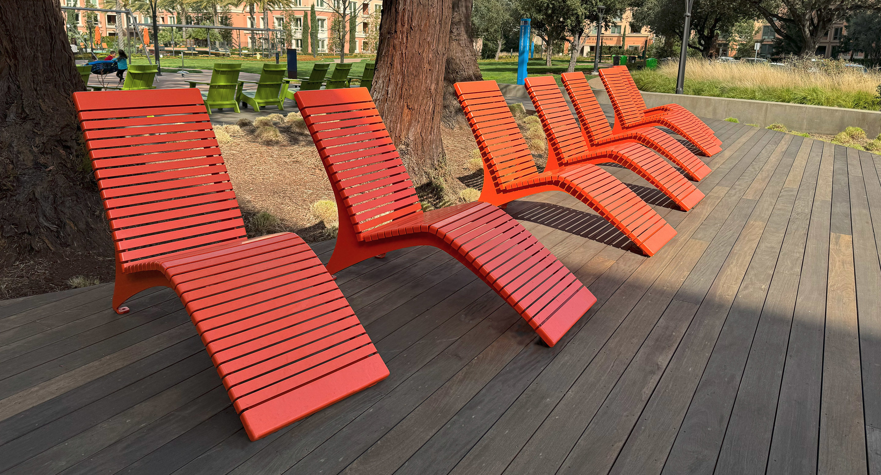 Six red slatted lounge chairs are arranged in a row on a wooden deck, with trees and green chairs visible in the background.