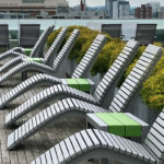 A row of modern white lounge chairs with green side tables on a wooden rooftop deck, with city buildings in the background.