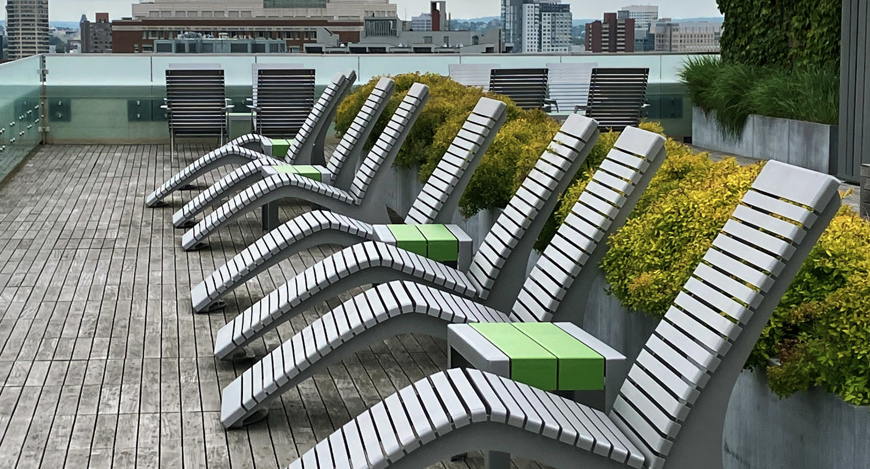 A row of modern white lounge chairs with green side tables on a wooden rooftop deck, with city buildings in the background.