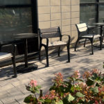 Quatre chaises de terrasse vides et deux petites tables sont disposées sur un trottoir pavé à l'extérieur d'un bâtiment, avec des fleurs au premier plan.
