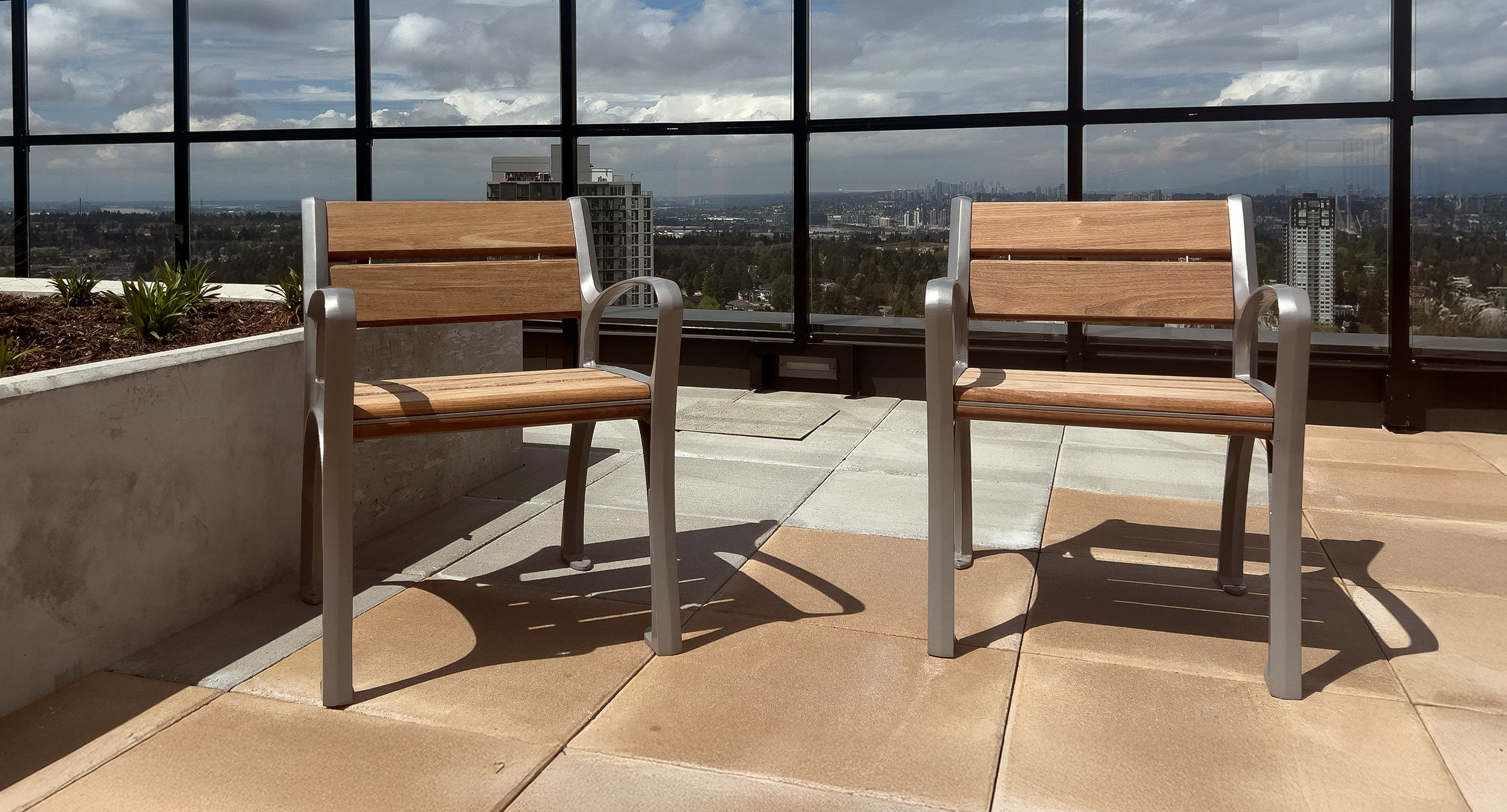 Deux chaises en bois avec des cadres en métal sont placées sur un patio sur le toit, avec des bâtiments urbains et un ciel nuageux visibles à l'arrière-plan à travers des panneaux de verre.