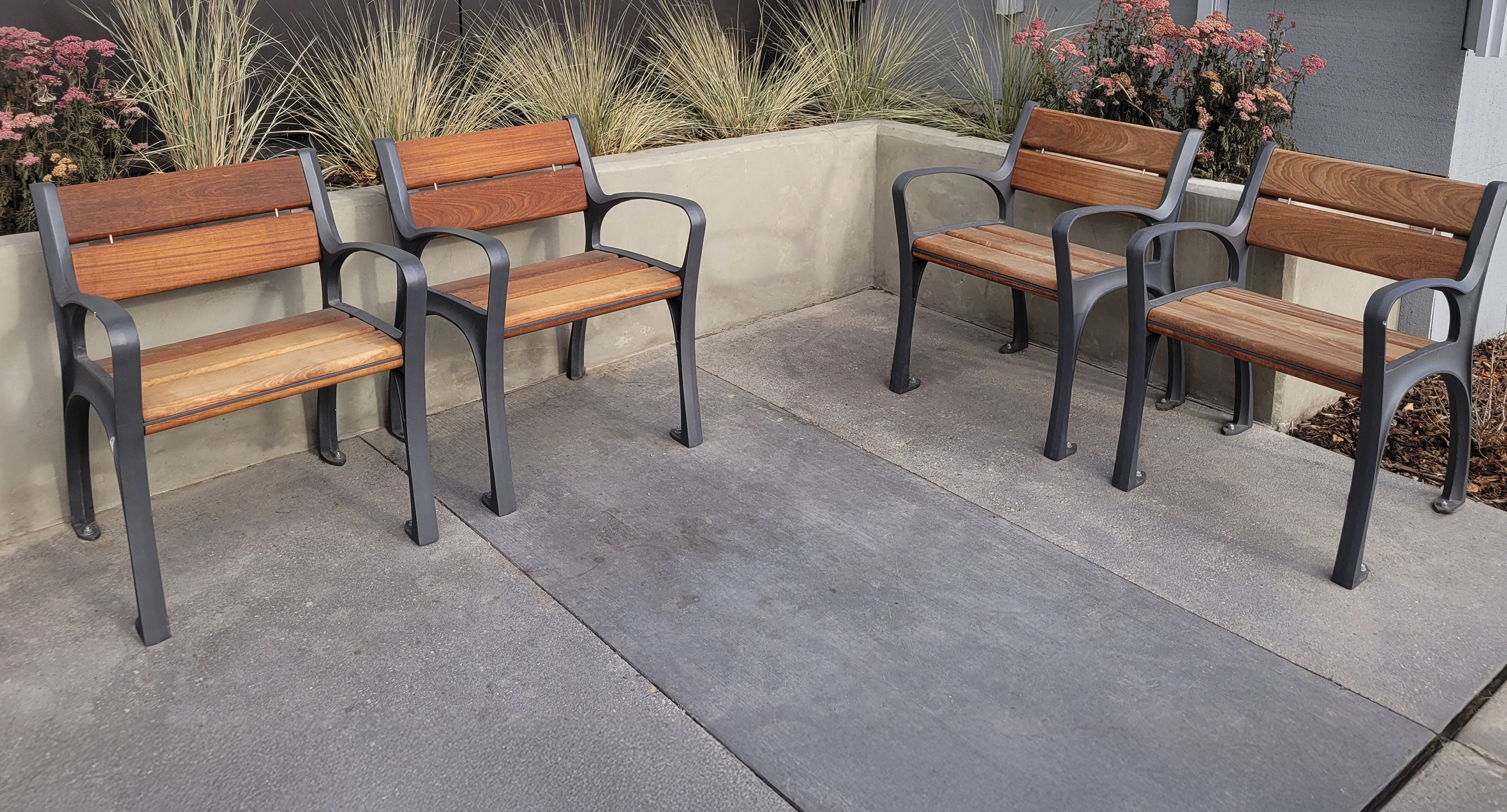 maglin_mch-0870-chairs-3 Quatre chaises en bois et en métal sont disposées en deux paires face à face sur un patio en béton, avec des jardinières et des graminées ornementales en arrière-plan.