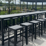 Long rectangular outdoor table with matching high stools on a paved patio, surrounded by greenery and additional seating areas.