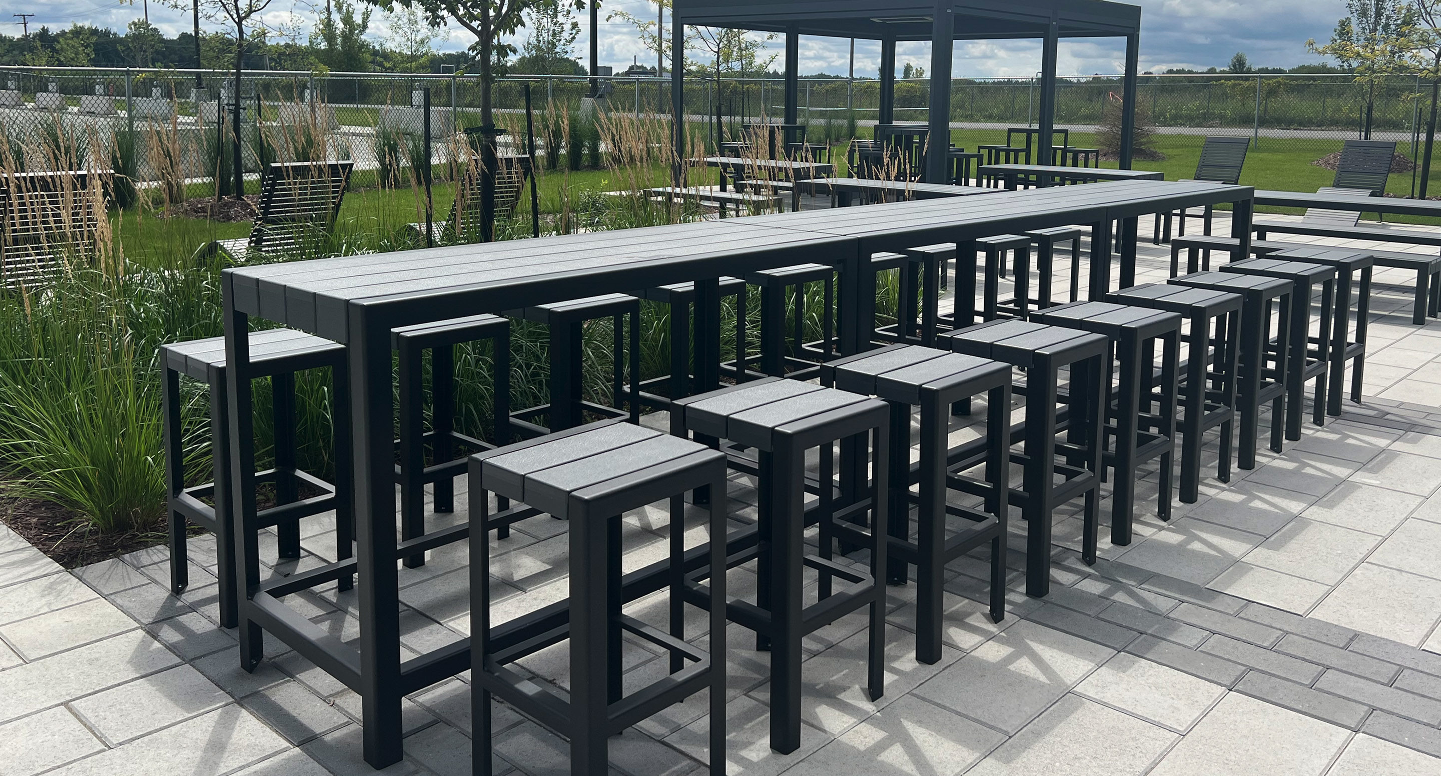 maglin_mch-1050-bar-height-stools-1 Long rectangular outdoor table with matching high stools on a paved patio, surrounded by greenery and additional seating areas.